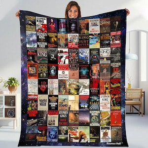 May include: A large, rectangular blanket featuring a collage of Stephen King book covers. The blanket has a dark blue background with a galaxy design. The book covers are arranged in a grid pattern, showcasing various titles and cover art.