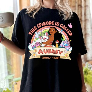 May include: Black t-shirt featuring a colorful graphic of a cartoon character with a rainbow, pig, and cake. The text reads "THIS EPISODE IS CALLED AUBREY TURNS TWO!" in a playful font. The shirt is a casual style.