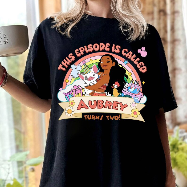 May include: Black t-shirt featuring a colorful graphic of a cartoon character with a rainbow, pig, and cake. The text reads "THIS EPISODE IS CALLED AUBREY TURNS TWO!" in a playful font. The shirt is a casual style.
