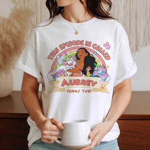 May include: White t-shirt featuring a colorful graphic with the text "THIS EPISODE IS CALLED AUBREY TURNS TWO!" and a cartoon illustration of a character with a pig, a chicken, and a cake. The design includes a rainbow and flowers.