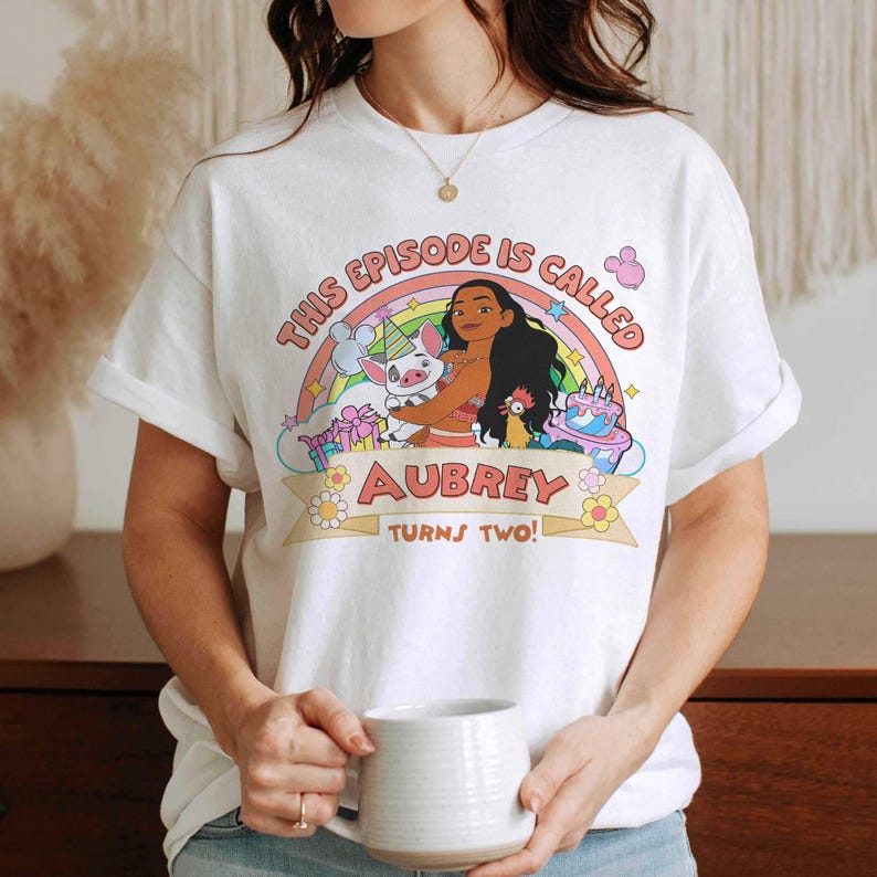 May include: White t-shirt featuring a colorful graphic with the text "THIS EPISODE IS CALLED AUBREY TURNS TWO!" and a cartoon illustration of a character with a pig, a chicken, and a cake. The design includes a rainbow and flowers.