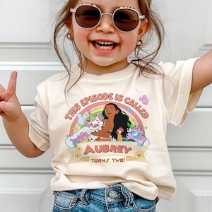 May include: A cream-colored t-shirt featuring a colorful graphic of a cartoon character, a pig, and a rooster, with a rainbow and a birthday cake. The text "THIS EPISODE IS CALLED" and "AUBREY TURNS TWO!" is printed on the shirt.