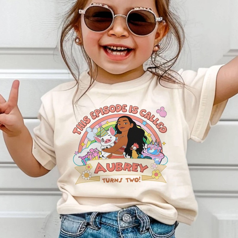 May include: A cream-colored t-shirt featuring a colorful graphic of a cartoon character, a pig, and a rooster, with a rainbow and a birthday cake. The text "THIS EPISODE IS CALLED" and "AUBREY TURNS TWO!" is printed on the shirt.