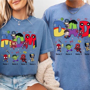 May include: Two blue t-shirts with superhero-themed designs. One shirt says "MOM" with various characters and the other says "DAD". Both shirts have space for names below the characters. The shirts are a light blue color.