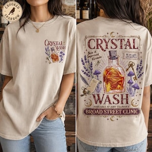 May include: A beige t-shirt featuring a small graphic on the front and a large graphic on the back. The back design displays a glass bottle filled with amber liquid, lavender sprigs, mushrooms, a key, and text reading Crystal Wash, New Improved Formula, and Broad Street Clinic.