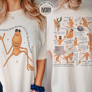 May include: Ivory-colored t-shirt featuring a cartoonish, orange creature with long limbs and the text "Big Badinki Bones shanked me in the leg at Panera Bread." The back of the shirt displays various cartoon illustrations and phrases.