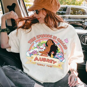 May include: Cream-colored t-shirt with a colorful graphic featuring a cartoon character, pig, and chicken. Text reads "THIS EPISODE IS CALLED AUBREY TURNS TWO!" above a rainbow and birthday cake. The shirt is worn by a person.