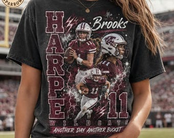 Op de afbeelding: Een antracietgrijs t-shirt met een vintage-stijl afbeelding van American footballspelers in bordeauxrode uniformen. Het ontwerp bevat de tekst "HARPE", "Brooks", "11", "WILDCATS" en "ANOTHER DAY ANOTHER BUCKET" met bliksemschichtaccenten.