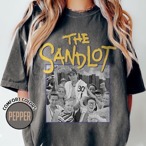 May include: A dark gray t-shirt featuring the movie title "The Sandlot" in yellow lettering above a black and white image of the movie cast. The shirt also has a "Comfort Colors" logo in a circle with the word "Pepper".