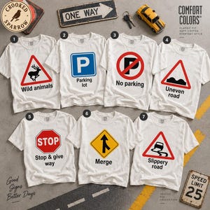 May include: Seven white t-shirts featuring various traffic sign graphics. The designs include a deer for wild animals, a P for parking lot, a no parking symbol, an uneven road sign, a stop sign, a merge sign, and a slippery road sign.