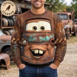 May include: A brown long-sleeve sweatshirt featuring a graphic of a rusty tow truck with large eyes, a wide smiling mouth with two prominent front teeth, and blue paint patches. The sleeves display tire tread patterns and text.