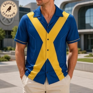 May include: A short-sleeve button-down shirt featuring a bold yellow X-shaped pattern across a deep blue background. The shirt has a notched collar and a buttoned front placket. A circular logo in the top left corner displays a bird and the text Crooked Sparrow.