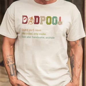 May include: A light beige t-shirt with the word "DADPOOL" in colorful letters. The text includes a definition: "like a dad, only cooler." The shirt also has the words "See also: handsome, asshole."