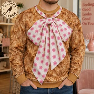 May include: A long-sleeved sweatshirt featuring a brown faux fur texture print and a large, attached light-colored neck scarf with a repeating pink floral pattern tied in a prominent bow at the center of the neckline.
