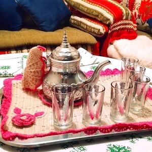 May include: A silver tea set with a teapot, six glasses, and a decorative tray. The glasses have a feather design. The tray is decorated with a pink crocheted border and a matching coaster. Pillows are in the background.