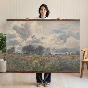 May include: A large landscape painting in a wooden frame. The artwork shows a field with trees beneath a cloudy sky. The colour palette includes blues, greens, and browns. The painting is held up by a person.