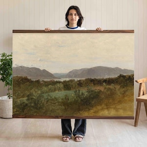 May include: A large landscape painting with a wooden frame. The artwork depicts a serene scene of mountains, a lake, and lush greenery. The color palette is dominated by earthy tones, creating a vintage aesthetic. The painting is held up by a person.