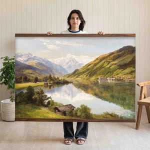 以下が含まれることがあります： 背景に山々が描かれた、穏やかな湖の風景を描いた額入りの風景画。 このアートワークは木製のフレームが特徴で、緑豊かな緑、雪をかぶった山頂、反射する湖など、自然の詳細な描写が施されています。 絵は人が持っています。