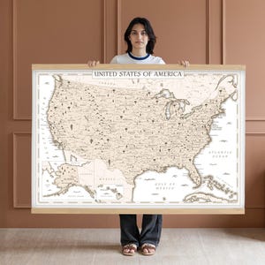 Pode incluir: Um grande mapa dos Estados Unidos da América, com uma estética vintage. O mapa é bege e castanho, com as fronteiras do país e as principais cidades rotuladas. O mapa é segurado por uma pessoa.