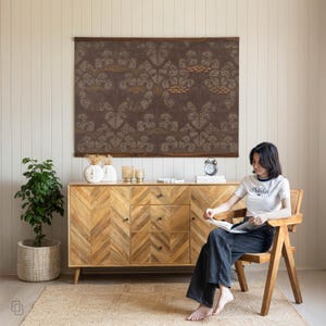 K&ouml;nnte beinhalten: Wohnzimmer mit einem Holzschrank mit Fischgr&auml;tmuster, einem gro&szlig;en gerahmten Kunstwerk mit braunem Blumendesign und einer Frau, die auf einem Holzstuhl sitzt. Eine Topfpflanze und ein gewebter Teppich vervollst&auml;ndigen die Einrichtung.