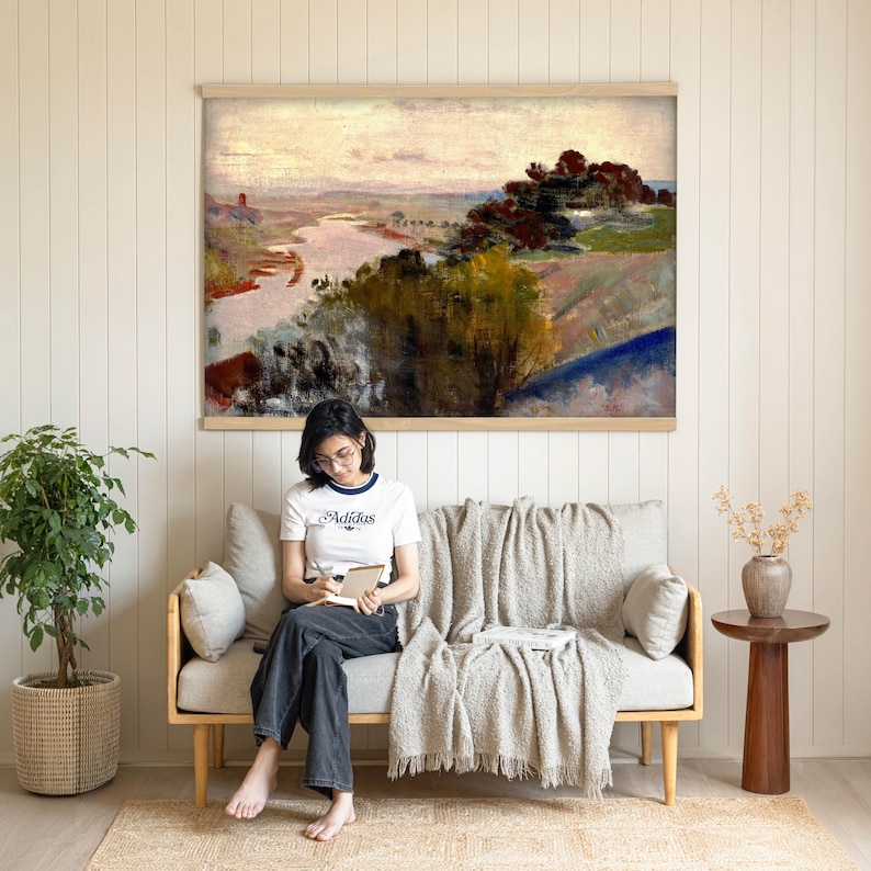 May include: A large landscape wall art print featuring an impressionist painting of a river winding through a hilly countryside with trees in shades of green, brown, and red under a soft, pale sky, displayed in a light wood frame.