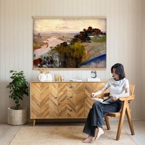 May include: A landscape painting in a light wood frame depicting a river winding through a hilly terrain with dark green trees and a soft, hazy sky. The artwork features muted tones of brown, green, and pale pink, capturing a serene outdoor scene.
