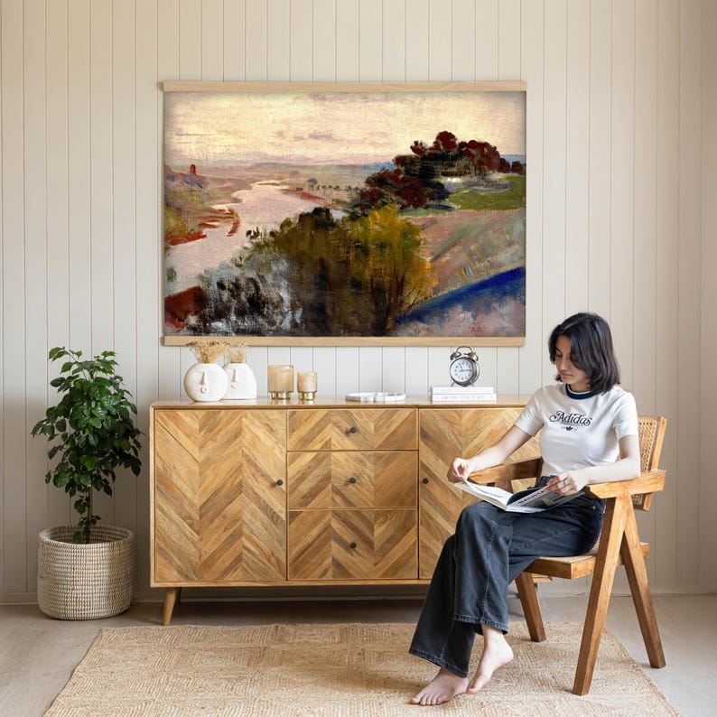 May include: A landscape painting in a light wood frame depicting a river winding through a hilly terrain with dark green trees and a soft, hazy sky. The artwork features muted tones of brown, green, and pale pink, capturing a serene outdoor scene.
