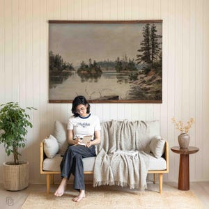 K&ouml;nnte beinhalten: Ein gro&szlig;es Landschaftsgem&auml;lde, das eine ruhige Seeszene mit B&auml;umen und Inseln zeigt, gerahmt in dunklem Holz. Das Kunstwerk h&auml;ngt an einer wei&szlig;en Wand &uuml;ber einem hellfarbigen Sofa. Eine Person mit einem wei&szlig;en T-Shirt und dunklen Jeans sitzt auf dem Sofa.