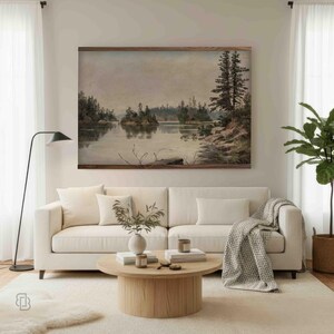 K&ouml;nnte beinhalten: Ein gerahmtes Landschaftsgem&auml;lde zeigt eine ruhige Seeszene mit Inseln und B&auml;umen. Das Kunstwerk in ged&auml;mpften T&ouml;nen h&auml;ngt &uuml;ber einem wei&szlig;en Sofa mit dekorativen Kissen und einer grauen Decke. Ein runder Couchtisch aus Holz und eine Stehlampe vervollst&auml;ndigen das Wohnzimmer.