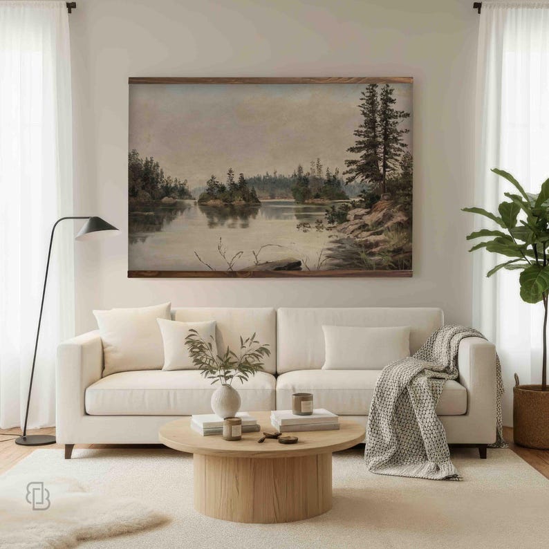 K&ouml;nnte beinhalten: Ein gerahmtes Landschaftsgem&auml;lde zeigt eine ruhige Seeszene mit Inseln und B&auml;umen. Das Kunstwerk in ged&auml;mpften T&ouml;nen h&auml;ngt &uuml;ber einem wei&szlig;en Sofa mit dekorativen Kissen und einer grauen Decke. Ein runder Couchtisch aus Holz und eine Stehlampe vervollst&auml;ndigen das Wohnzimmer.