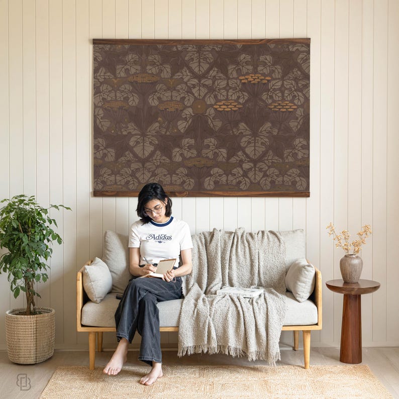 K&ouml;nnte beinhalten: Ein gro&szlig;er, rechteckiger Wandbehang mit braunem Hintergrund und einem sich wiederholenden Muster aus stilisierten Bl&auml;ttern und Blumen in Grau- und Beiget&ouml;nen. Der Wandbehang ist auf einem Holzrahmen montiert. Das Bild zeigt eine Wohnzimmereinrichtung mit Sofa, Teppich und Topfpflanze.