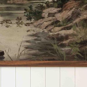 K&ouml;nnte beinhalten: Ein Vintage-Landschaftsgem&auml;lde, das eine ruhige Wasserlandschaft mit felsiger K&uuml;ste und &uuml;ppigem Gr&uuml;n darstellt. Das Kunstwerk hat einen Holzrahmen und ist an einer wei&szlig;en Wand mit Paneelen befestigt, was eine rustikale &Auml;sthetik erzeugt.