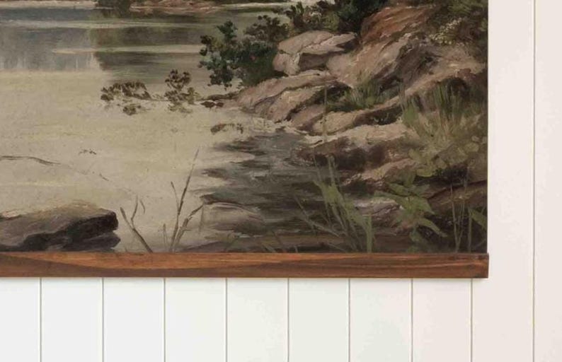 K&ouml;nnte beinhalten: Ein Vintage-Landschaftsgem&auml;lde, das eine ruhige Wasserlandschaft mit felsiger K&uuml;ste und &uuml;ppigem Gr&uuml;n darstellt. Das Kunstwerk hat einen Holzrahmen und ist an einer wei&szlig;en Wand mit Paneelen befestigt, was eine rustikale &Auml;sthetik erzeugt.