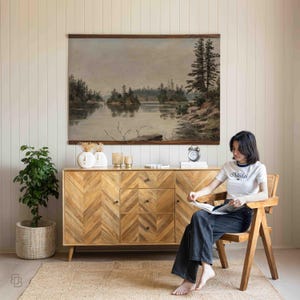 K&ouml;nnte beinhalten: Ein Holzsideboard mit Fischgr&auml;tmuster, gekr&ouml;nt von dekorativen Vasen, Kerzen und einer Uhr. Dar&uuml;ber h&auml;ngt ein gro&szlig;es Landschaftsgem&auml;lde. Eine Frau sitzt in einem Holzstuhl und liest ein Buch. Ein Topfpflanze und ein gewebter Teppich.