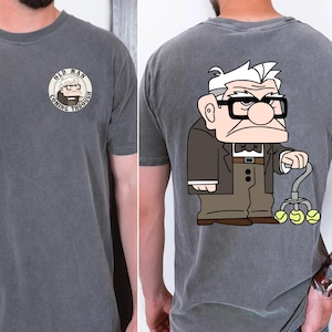 May include: Charcoal gray t-shirt featuring a cartoon illustration of an elderly man with glasses and a cane holding tennis balls. The front of the shirt has a circular graphic with the text "OLD MAN COMING THROUGH".