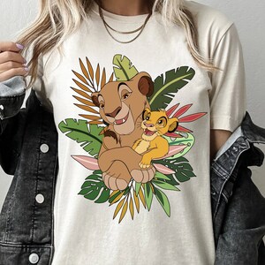 May include: A beige t-shirt with a cartoon image of Simba and Mufasa from The Lion King. The image features the two lions in a jungle setting with green and brown leaves.