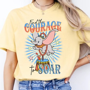 May include: A yellow t-shirt with a cartoon image of Dumbo the elephant flying through the air with a circus ring. The text "Find the Courage to SOAR" is printed on the shirt.
