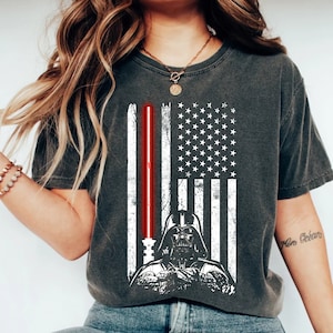 May include: A dark gray t-shirt with a distressed white American flag design. Darth Vader is standing in front of the flag, holding a red lightsaber. The text "More Ocean" is written on the right sleeve of the shirt.