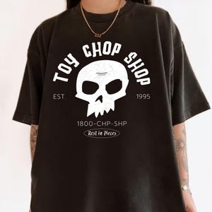 Pu&ograve; includere: Una t-shirt nera a maniche corte con una grafica bianca raffigurante un teschio e il testo TOY CHOP SHOP, EST. 1995, 1800-CHP-SHP e Rest in Pieces. Il design del teschio presenta una texture screpolata e vissuta, centrato sul davanti.
