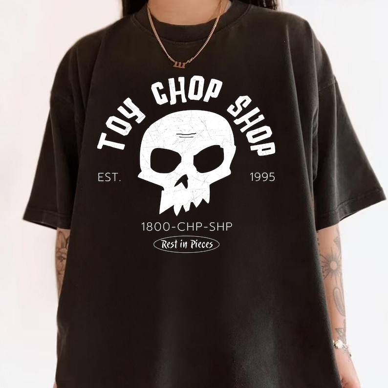 Pu&ograve; includere: Una t-shirt nera a maniche corte con una grafica bianca raffigurante un teschio e il testo TOY CHOP SHOP, EST. 1995, 1800-CHP-SHP e Rest in Pieces. Il design del teschio presenta una texture screpolata e vissuta, centrato sul davanti.