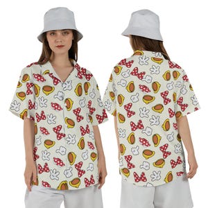 May include: Off-white button-down shirt with a repeating pattern of red polka-dot bows, yellow gloves, and yellow ovals. The shirt has short sleeves and a collared neckline. The model is wearing a white bucket hat and white shorts.