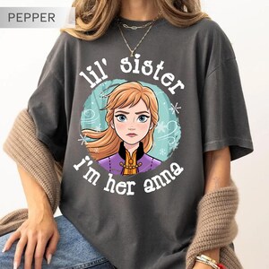 May include: Dark gray t-shirt featuring a cartoon image of a character with blonde hair and blue eyes, surrounded by a blue and white design. The text "lil' sister I'm her anna" is printed above and below the image.