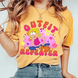 May include: A mustard yellow short-sleeve t-shirt featuring a graphic print of a cartoon character with blonde hair, a pink top, and blue pants. The text OUTFIT REPEATER is printed in pink and orange bubble letters above and below the character.