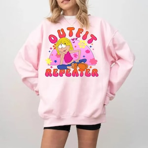 May include: A light pink crewneck sweatshirt featuring a graphic print of a cartoon character with blonde hair, a pink top, and blue pants. The text "OUTFIT REPEATER" is printed in bold, orange, bubble-style letters above and below the character.