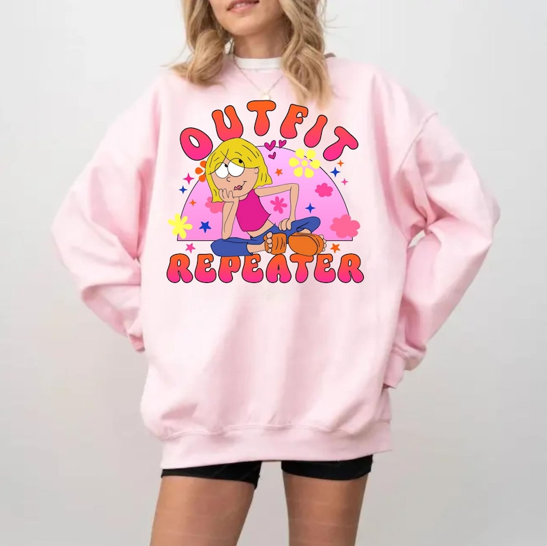 May include: A light pink crewneck sweatshirt featuring a graphic print of a cartoon character with blonde hair, a pink top, and blue pants. The text "OUTFIT REPEATER" is printed in bold, orange, bubble-style letters above and below the character.