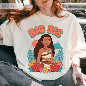 May include: Ivory-colored t-shirt with the text "BIG SIS" in a retro font. The graphic features a cartoon woman with long brown hair, a necklace, and a floral design. The design includes a sun, mountains, and tropical flowers.