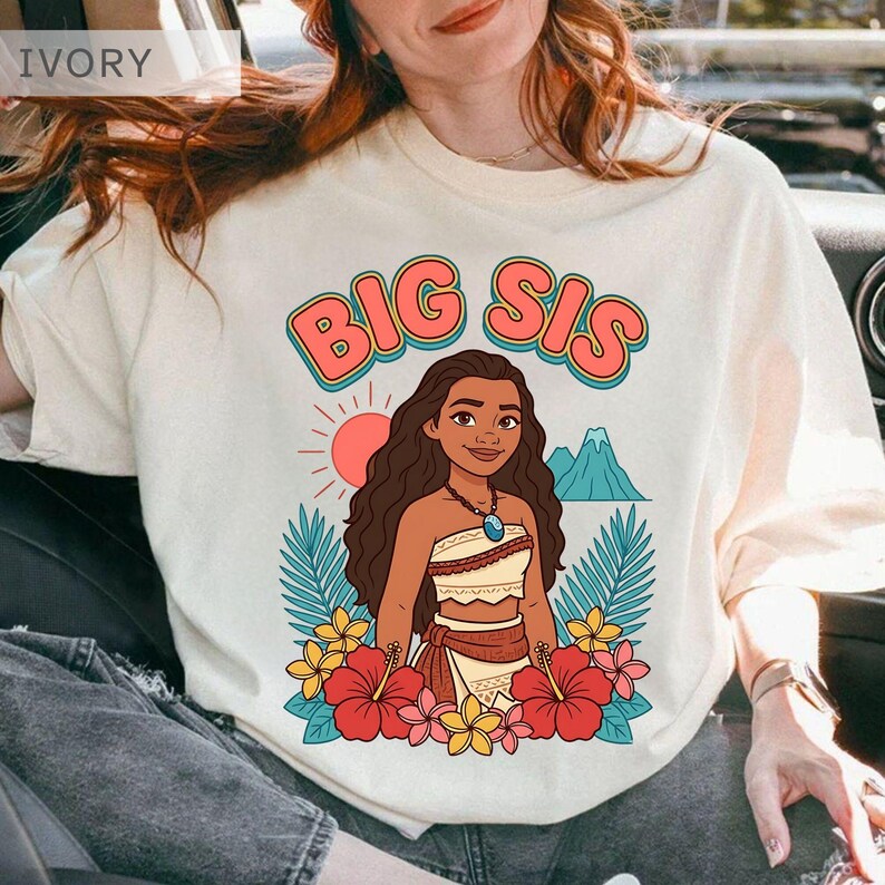 May include: Ivory-colored t-shirt with the text "BIG SIS" in a retro font. The graphic features a cartoon woman with long brown hair, a necklace, and a floral design. The design includes a sun, mountains, and tropical flowers.