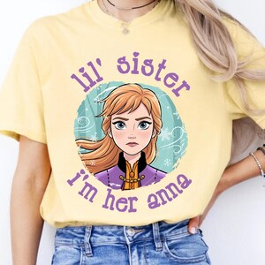 May include: Butter-colored t-shirt featuring a cartoon image of Anna from Frozen, surrounded by a teal circle with snowflakes. The text "lil' sister, I'm her anna" is printed in purple around the image. The shirt is worn with blue jeans.