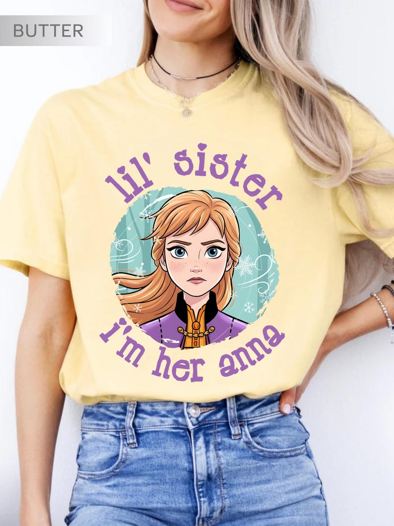 May include: Butter-colored t-shirt featuring a cartoon image of Anna from Frozen, surrounded by a teal circle with snowflakes. The text "lil' sister, I'm her anna" is printed in purple around the image. The shirt is worn with blue jeans.