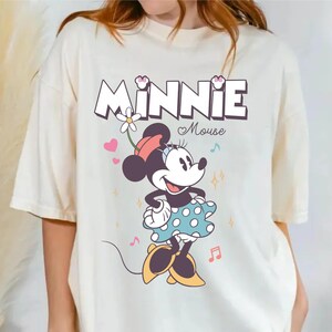 Puede incluir: Una camiseta blanca con un gr&aacute;fico de Minnie Mouse con un vestido azul de lunares y un lazo rojo. El texto "Minnie Mouse" est&aacute; impreso encima de ella en una fuente colorida.